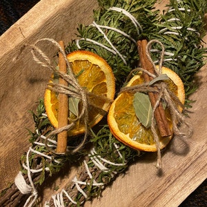 May include: Two bundles of green sage tied with white twine, adorned with dried orange slices, cinnamon sticks, and bay leaves. The bundles rest in a wooden bowl, creating a rustic and natural aesthetic. The sage bundles are a natural home decor item.