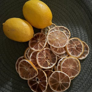 May include: Two fresh yellow lemons and a pile of dried lemon slices on a dark green surface. The dried slices are a light brown colour with a darker brown centre. The lemons have a textured skin.