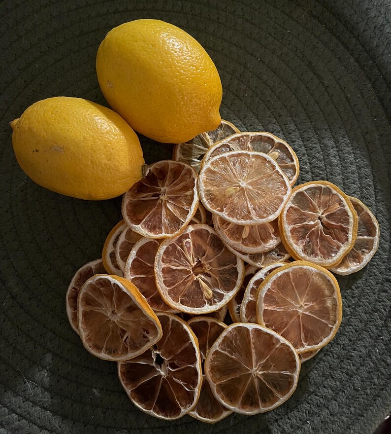 May include: Two fresh yellow lemons and a pile of dried lemon slices on a dark green surface. The dried slices are a light brown colour with a darker brown centre. The lemons have a textured skin.