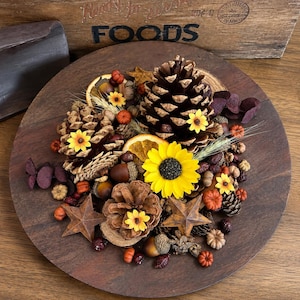 May include: A decorative arrangement featuring brown pinecones, dried orange slices, small yellow sunflowers, acorns, and miniature orange pumpkins. Two rusted metal stars are placed among the natural elements on a circular dark wood base.