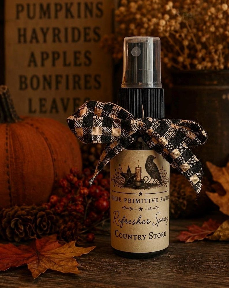 May include: A clear plastic spray bottle with a black pump top, adorned with a black and tan buffalo check ribbon. The label features a black crow, a star, a candle, and the text: OLDE PRIMITIVE FARM Refresher Spray COUNTRY STORE.