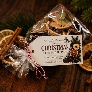 May include: A clear plastic bag filled with dried fruit, cinnamon sticks, and evergreen sprigs, tied with red and white twine. A cream-colored tag attached to the bag features the text "OLDE PRIMITIVE FARM CHRISTMAS SIMMER POT" and illustrations.