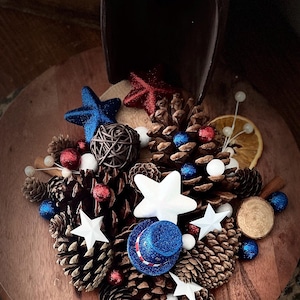 May include: A patriotic arrangement featuring pine cones, white and blue stars, red and blue glitter balls, and a miniature blue top hat. The arrangement is set on a wooden surface, with a dark wooden scoop in the background.
