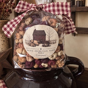 May include: A clear plastic bag filled with small, multi-colored fabric pumpkins, tied with a red and white gingham ribbon. A circular label features a house illustration and the text: OLDE PRIMITIVE FARM APPLE CINNAMON HANDMADE IN RI.