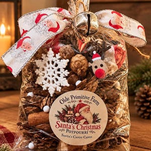 May include: A clear plastic bag filled with pinecones, cinnamon sticks, and small white pom-poms. It features a white snowflake ornament, a wooden reindeer, and a large silver jingle bell. A ribbon with Santa Claus prints and a label reading Olde Primitive Farm Santa's Christmas Potpourri Santa's Candy Canes are attached.