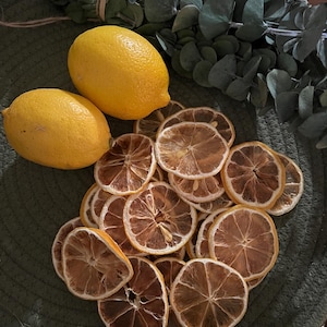 May include: Two fresh yellow lemons and a pile of dried lemon slices are arranged on a green textured surface. Eucalyptus leaves are in the background. The dried lemon slices have a brown, textured appearance.