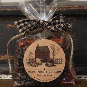 May include: A clear plastic bag filled with dark brown and orange potpourri, tied with a black and tan buffalo check ribbon. A round tan label features a drawing of a red house, a sheep, and a doll, with text reading OLDE PRIMITIVE FARM.
