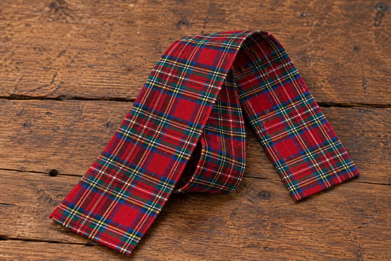 May include: A long, rectangular fabric strip featuring a red, green, blue, and yellow plaid tartan pattern. The textile is folded into a loop, displaying the consistent checkered design across its entire surface with finished edges.