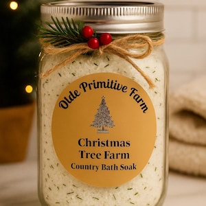 May include: A clear glass jar filled with white bath soak, labeled "Olde Primitive Farm Christmas Tree Farm Country Bath Soak." The jar is adorned with a twine bow, sprig of evergreen, and red berries. The background includes a Christmas tree.