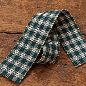 May include: A length of fabric ribbon featuring a dark forest green and cream white plaid pattern. The ribbon is folded into a loose loop, displaying the woven texture of the material and the consistent grid design across the entire surface.