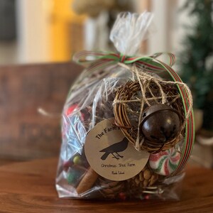 May include: A clear plastic bag filled with a variety of Christmas decorations, including a small wreath with a bell, pine cones, cinnamon sticks, and candy canes. The bag is tied with a red and green ribbon and has a tag that reads "The Farm Christmas Tree Farm Rhode Island".