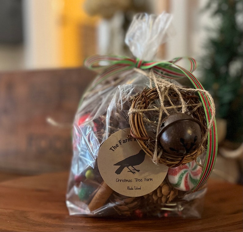 May include: A clear plastic bag filled with a variety of Christmas decorations, including a small wreath with a bell, pine cones, cinnamon sticks, and candy canes. The bag is tied with a red and green ribbon and has a tag that reads "The Farm Christmas Tree Farm Rhode Island".