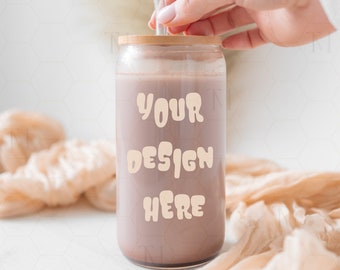 May include: A clear glass can-shaped tumbler with a wooden lid and a clear straw. The tumbler is filled with a light brown liquid and has the words "YOUR DESIGN HERE" printed on the front in a beige color. The tumbler is on a white surface.