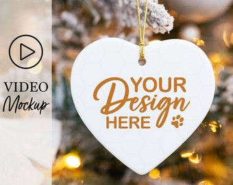 May include: White ceramic heart ornament with a gold string hanging from a snowy Christmas tree. The ornament has the text "YOUR Design HERE" in brown lettering.