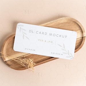 May include: A white DL card mockup with text and botanical illustrations, measuring 8.27 x 3.9 inches, rests on a wooden oval tray. The tray is light brown, and a small bundle of dried flowers is placed near the card.
