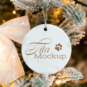 K&ouml;nnte beinhalten: Wei&szlig;er Keramik-Weihnachtsbaumschmuck mit einem braunen Pfotenabdruck und dem Text "tita mockup", der an einem schneebedeckten Weihnachtsbaum h&auml;ngt.