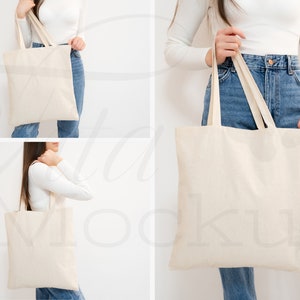 May include: A person is holding a white canvas tote bag with two handles. The bag is empty and has a natural, unbleached look. The person is wearing blue jeans and a white long-sleeved shirt.