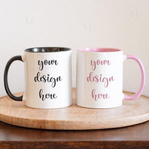 May include: Two white ceramic mugs with black and pink rims. The mugs have the text "your design here" written in black cursive on them. The mugs are sitting on a wooden coaster.