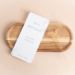 Puede incluir: Una maqueta de menú blanca con el texto «MENU MOCKUP» y dimensiones de 9,9 x 21 cm, sobre una bandeja ovalada de madera. Flores secas colocadas en la bandeja, con un fondo rosa claro.