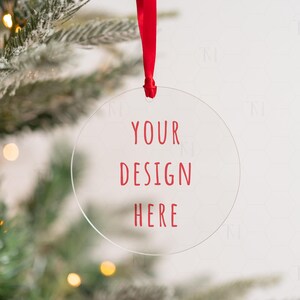 May include: A clear round ornament with a red ribbon hanging from a Christmas tree. The ornament has the text "YOUR DESIGN HERE" printed in red.