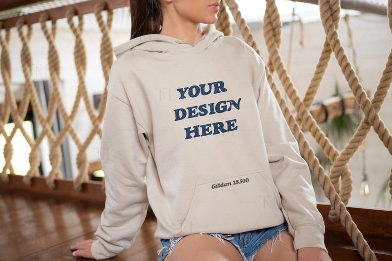 Op de afbeelding: Een beige hoodie met een blauwe print die "YOUR DESIGN HERE" en "Gildan 18500" in de rechterbenedenhoek zegt.