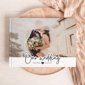 May include: A white wedding photo album with a photo of a bride and groom kissing under a veil. The album has the text "Our wedding" in black cursive writing and the names "Olivia Wilson" and "Aaron Loeb" below it.