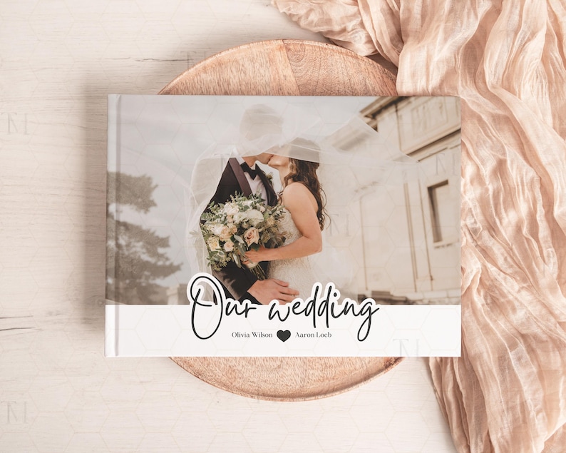 May include: A white wedding photo album with a photo of a bride and groom kissing under a veil. The album has the text "Our wedding" in black cursive writing and the names "Olivia Wilson" and "Aaron Loeb" below it.