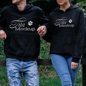 May include: Two black hoodies with the text "Tita Mockup" and a paw print design. The hoodies have a front pocket and a hood. The people are wearing blue jeans and standing next to a wooden fence.