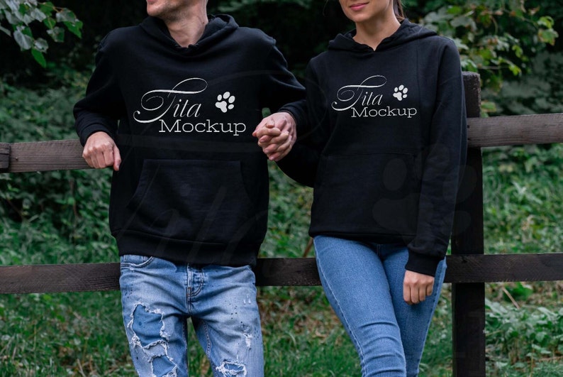 May include: Two black hoodies with the text "Tita Mockup" and a paw print design. The hoodies have a front pocket and a hood. The people are wearing blue jeans and standing next to a wooden fence.