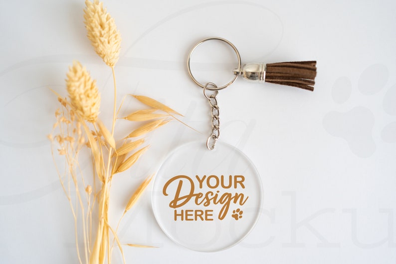 May include: A clear, round acrylic keychain with the text "YOUR DESIGN HERE" in a gold script font, featuring a paw print. The keychain has a silver ring, chain, and a brown tassel. Dried wheat stalks are arranged on the left side.