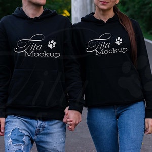 May include: Two people wearing black hoodies with white text that says "Jita Mockup" and a paw print. They are walking down a paved road with green trees on either side.