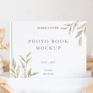 May include: A white photo book mockup with a watercolor design of green leaves on the cover. The book is 11 x 8 inches and is labeled "Hard Cover" and "Photo Book Mockup".