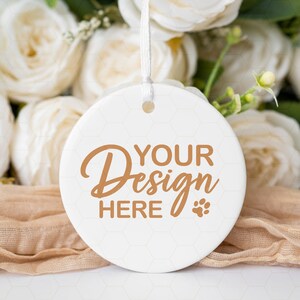 May include: White ceramic ornament with a brown inscription that reads "Your Design Here" and a small paw print. The ornament is hanging from a white ribbon and is surrounded by white roses and a beige fabric.