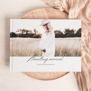 May include: A white photo album with a rustic wood background. The album cover features a photo of a pregnant woman in a field, wearing a white dress and a wide-brimmed hat. The text "Awaiting arrival" and "Our precious 9 months" is printed on the album cover.