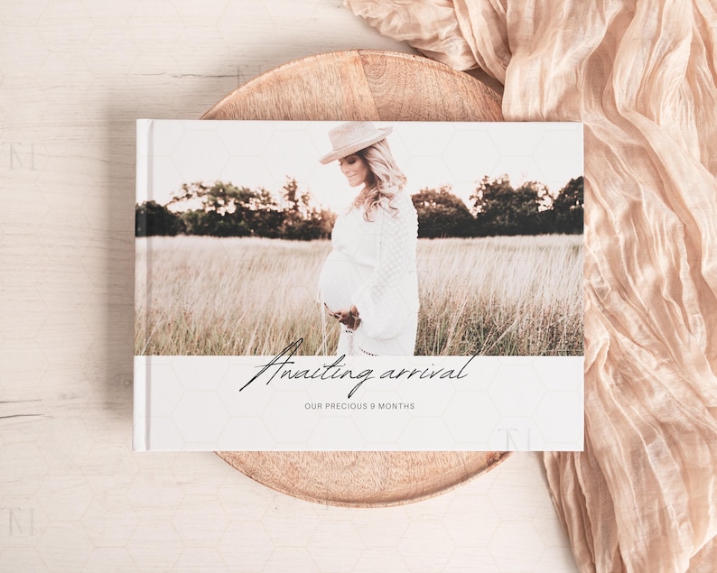 May include: A white photo album with a rustic wood background. The album cover features a photo of a pregnant woman in a field, wearing a white dress and a wide-brimmed hat. The text "Awaiting arrival" and "Our precious 9 months" is printed on the album cover.