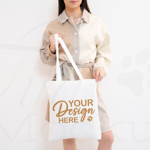 Può includere: Una borsa tote bianca con il testo "YOUR Design HERE" e un'impronta di zampa marrone. La borsa è tenuta da una persona che indossa una camicia beige e bianca.
