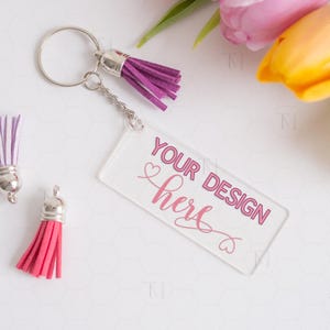 May include: A clear acrylic keychain with a pink heart and the text "YOUR DESIGN here" in pink. The keychain is attached to a silver key ring with a purple tassel.
