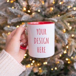 May include: A white ceramic mug with a red rim, featuring the text "YOUR DESIGN HERE" in red lettering. The mug is being held by a hand against a blurred background of a Christmas tree with white snow and twinkling lights.