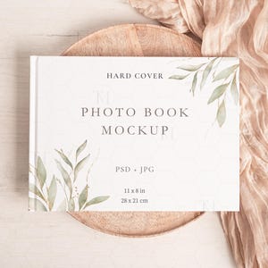 Op de afbeelding: Een witte fotoboek mockup met een harde kaft en een aquarelontwerp van groene bladeren. Het boek ligt op een houten dienblad en een beige stof is over het dienblad gedrapeerd. De tekst op het boek luidt "HARD COVER PHOTO BOOK MOCKUP PSD JPG 11 x 8 in 28 x 21 cm".