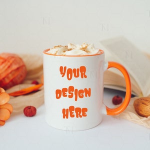 May include: White ceramic mug with an orange rim and handle. The mug has the text "YOUR DESIGN HERE" in orange lettering. The mug is filled with a creamy beverage topped with whipped cream and cinnamon.