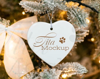 Könnte beinhalten: Weißer, herzförmiger Keramikanhänger mit dem Text "Tita Mockup" und einem braunen Pfotenabdruck. Der Anhänger hängt an einem schneebedeckten Weihnachtsbaumzweig.
