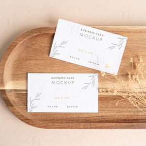 May include: Two white business card mockups with the text "BUSINESS CARD MOCKUP" and dimensions of 9 x 5 cm and 3.5 x 2 in. The cards are on a wooden tray with dried plants. The background is a light beige color.