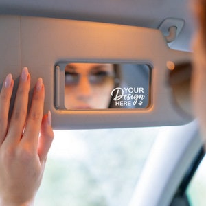 May include: A close-up of a car's sun visor with a mirror. The mirror reflects a person's face with the text "YOUR DESIGN HERE" written on it.