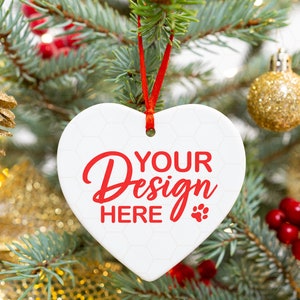 May include: White heart-shaped ceramic bauble with red ribbon hanging on a Christmas tree. The bauble has the text "YOUR Design HERE" in red lettering.