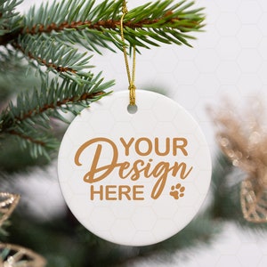 Op de afbeelding: Witte kerstbal van keramiek met een gouden koord dat aan een dennentak hangt. De kerstbal heeft de tekst "YOUR Design HERE" in gouden letters met een pootprint.