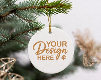 Peut inclure: Ornement de Noël en céramique blanche avec une ficelle dorée suspendue à une branche de pin. L'ornement porte le texte "YOUR Design HERE" en lettres dorées avec une empreinte de patte.