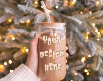 Puede incluir: Un vaso de vidrio transparente con tapa de madera y pajita transparente, lleno de líquido marrón. Las palabras "YOUR DESIGN HERE" están impresas en la parte delantera con una fuente retro. El fondo presenta un árbol de Navidad borroso con luces doradas.