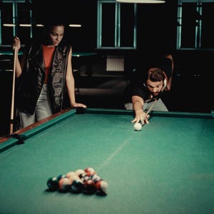 May include: A man and a woman playing snooker in a dimly lit room. The man is taking a shot, while the woman watches. There are several snooker balls on the green felt table.