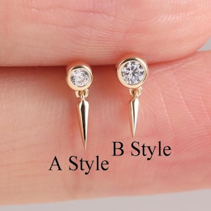 May include: Two gold-colored stud earrings with a small, round, clear gemstone and a dangling, pointed spike. The earrings are labeled "A Style" and "B Style" in black text. The background is a light pink color.