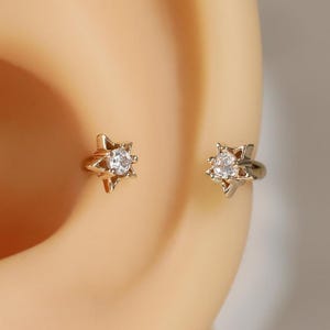 May include: Two gold-toned star-shaped earrings, each featuring a central clear, round-cut gemstone. The star designs have pointed edges and are positioned on the ear cartilage, showcasing a metallic finish and a sparkling stone center.